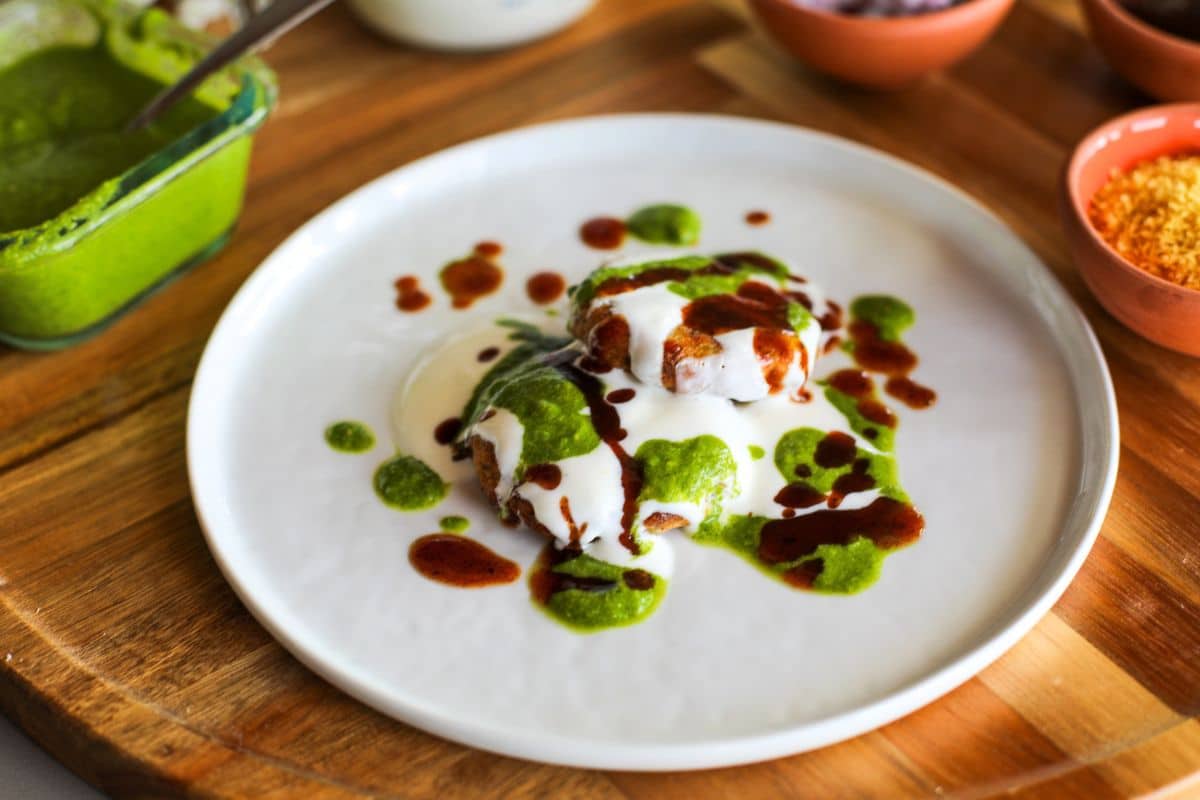 Two small potato patties topped with white yogurt, green chutney, and brown sauce are served on a white plate, with condiments in bowls nearby on a wooden table.