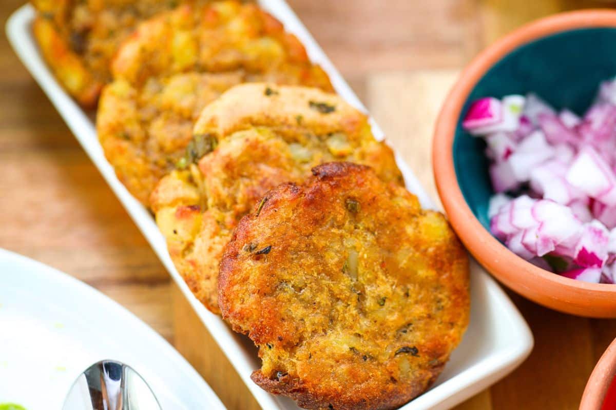 Several golden-brown potato patties are arranged on a rectangular white plate beside a bowl of chopped red onions.