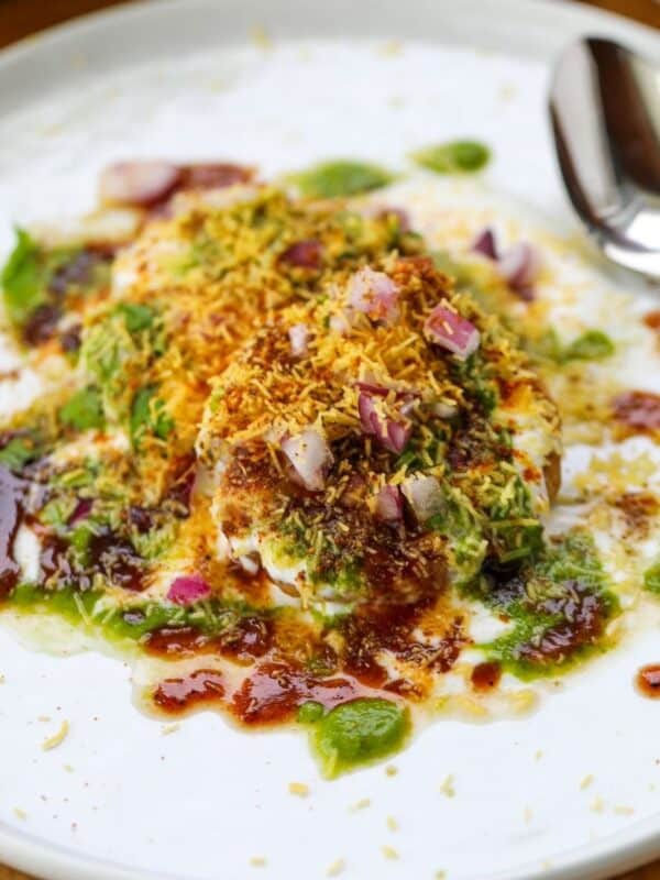 A plate of aloo tikki chaat topped with yogurt, chutneys, chopped onions, sev, and spices, with a spoon placed on the side of the dish.