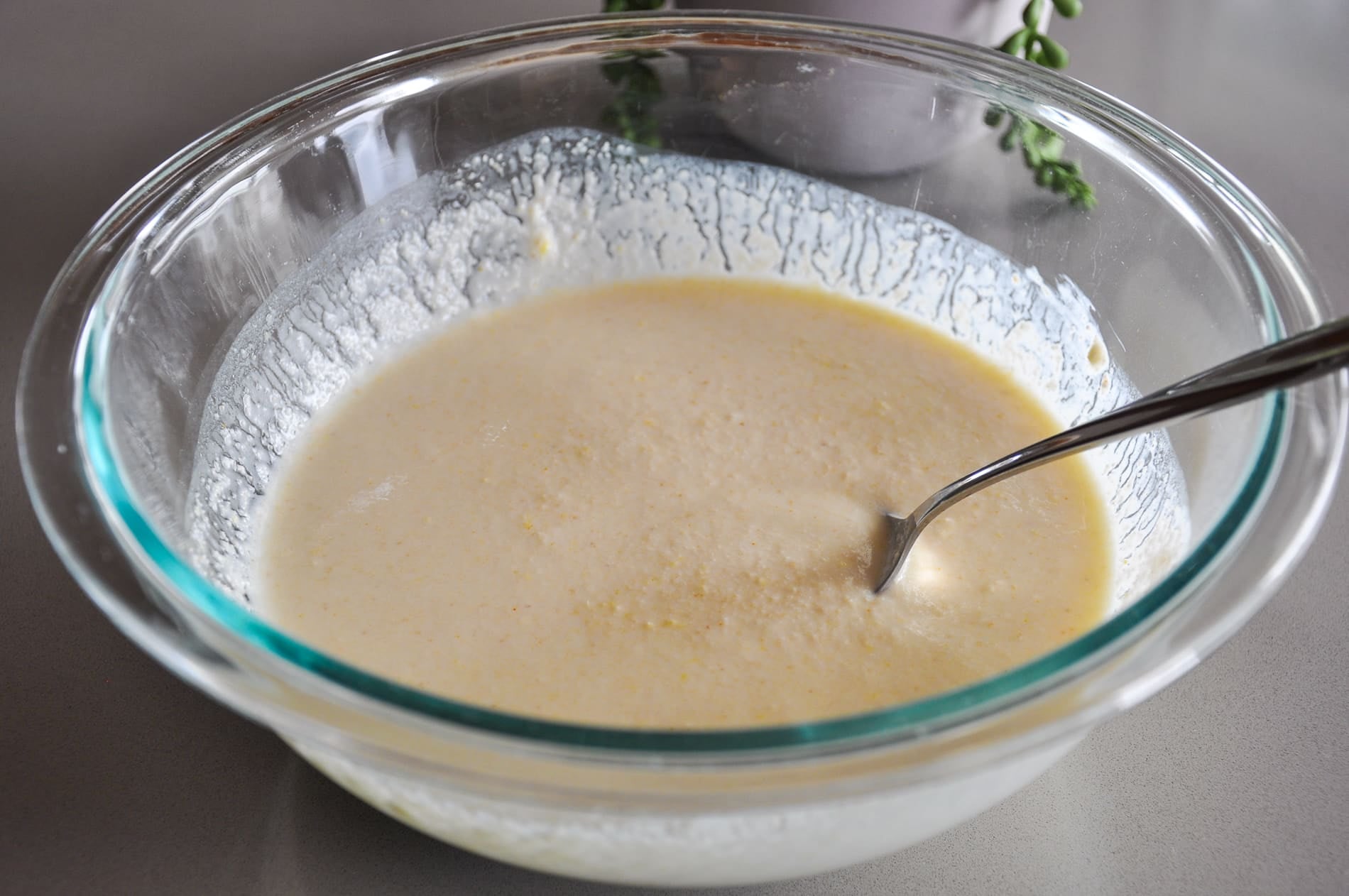 Clear glass bowl with pale yellow dhokla batter and a metal spoon inside, placed on a gray surface.