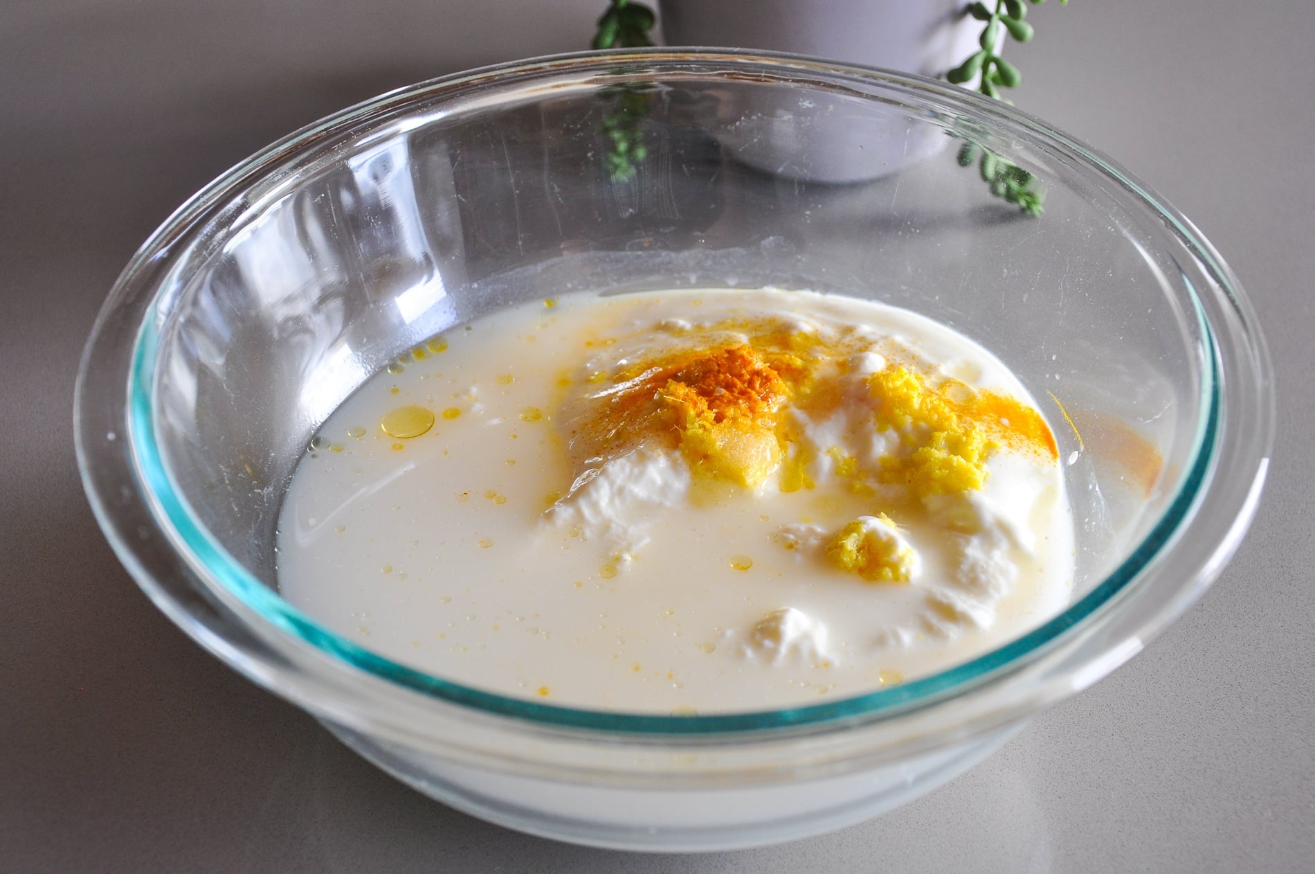 A glass bowl contains rava, water, yogurt, turmeric, and ginger, with small spots of oil on the surface.