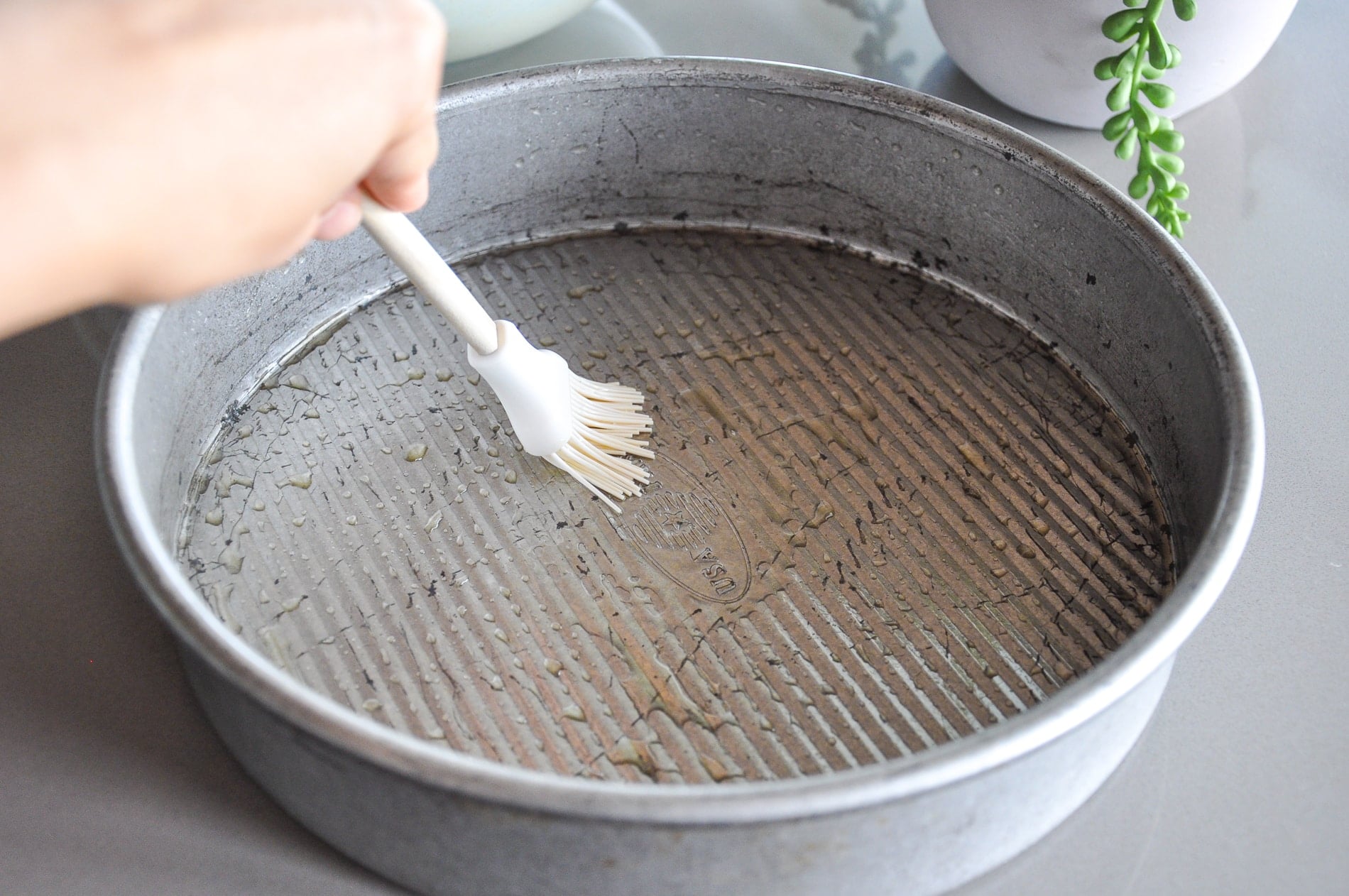 A hand uses a brush to apply oil inside a round metal baking pan, preparing it for baking.