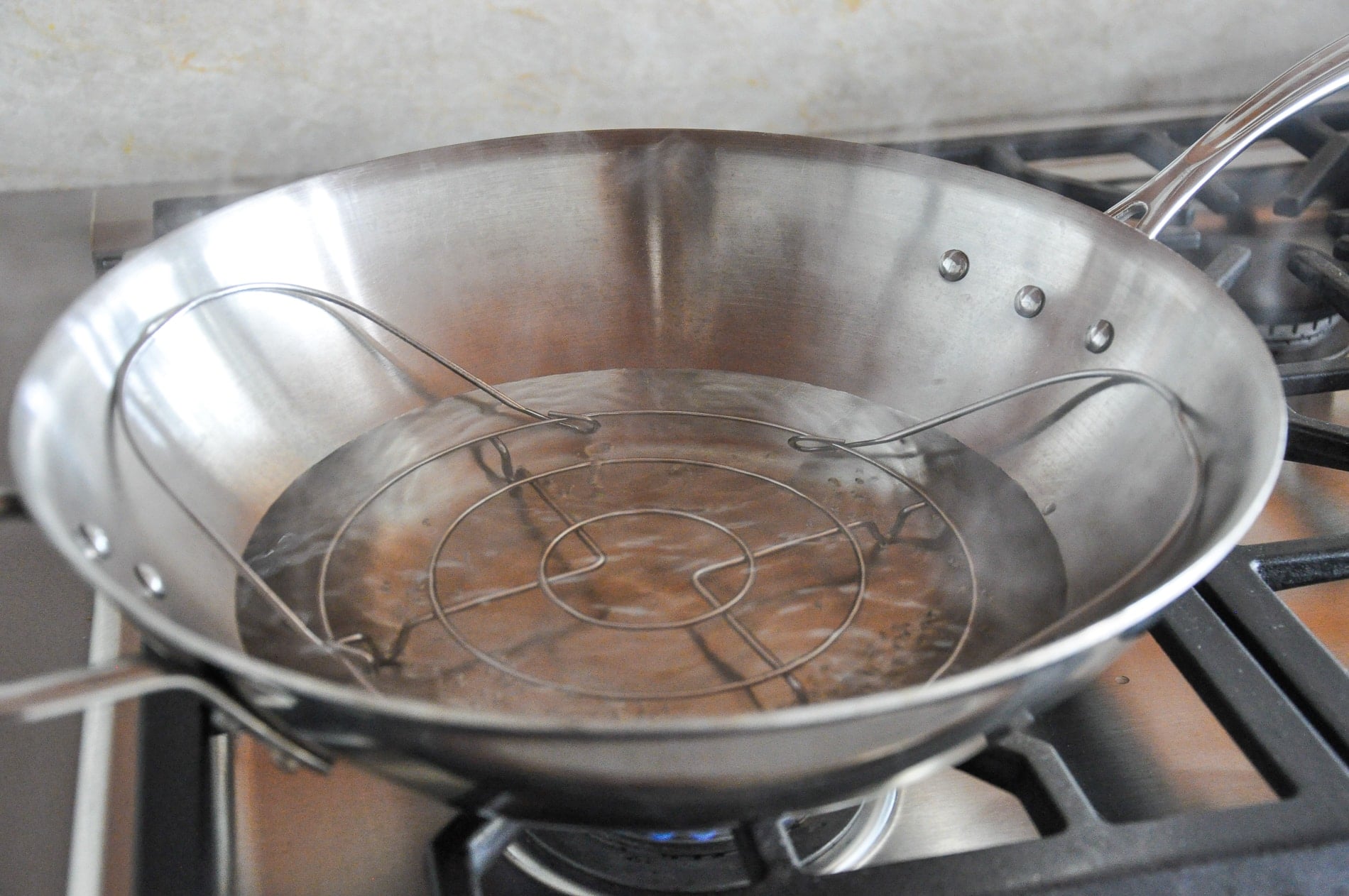 A stainless steel frying pan with a metal rack sits on a gas stove burner with water boiling inside.