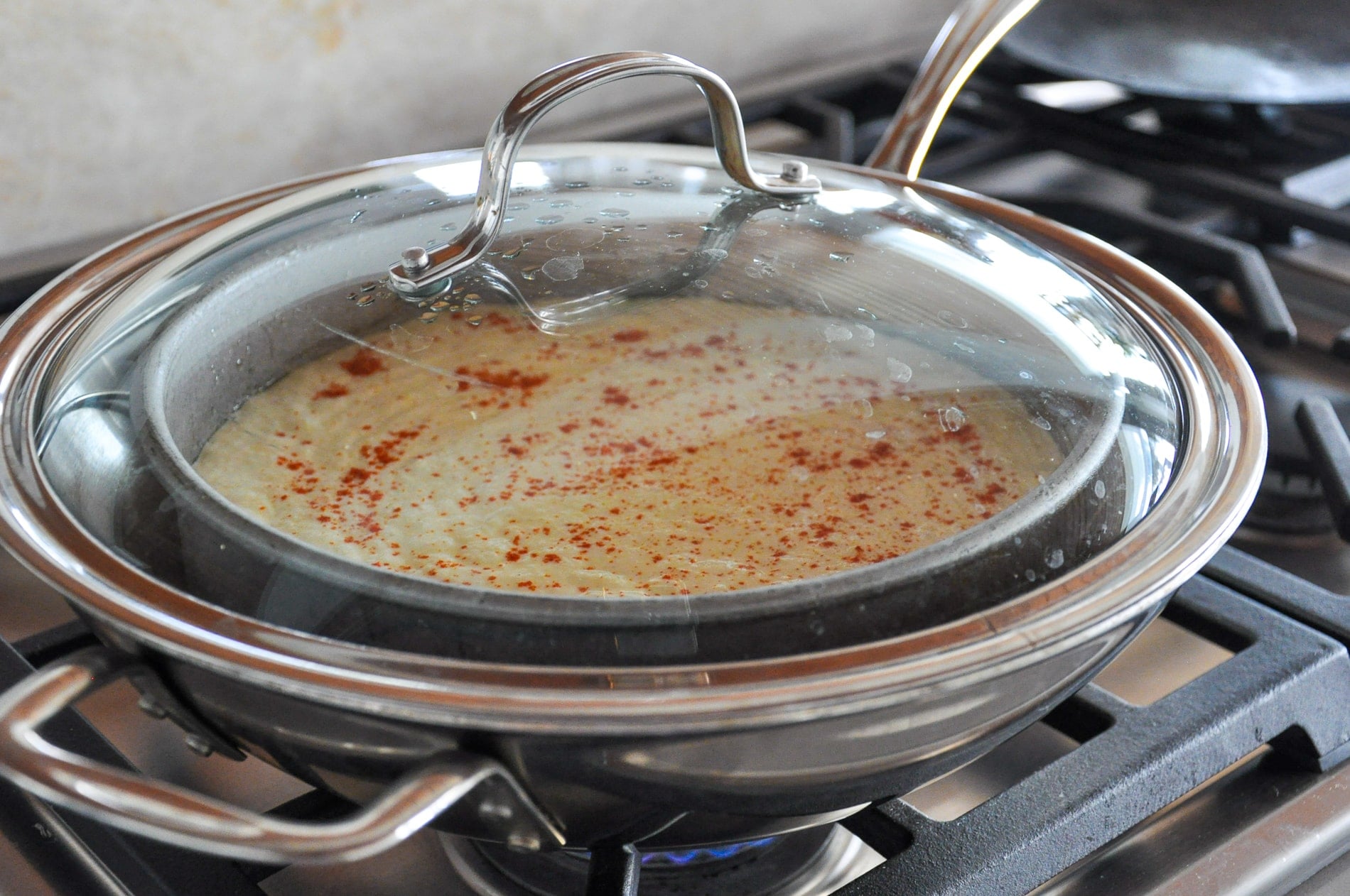 A covered skillet on a stovetop contains a cake pan containing dhokla batter sprinkled with red chili powder, cooking under a glass lid.