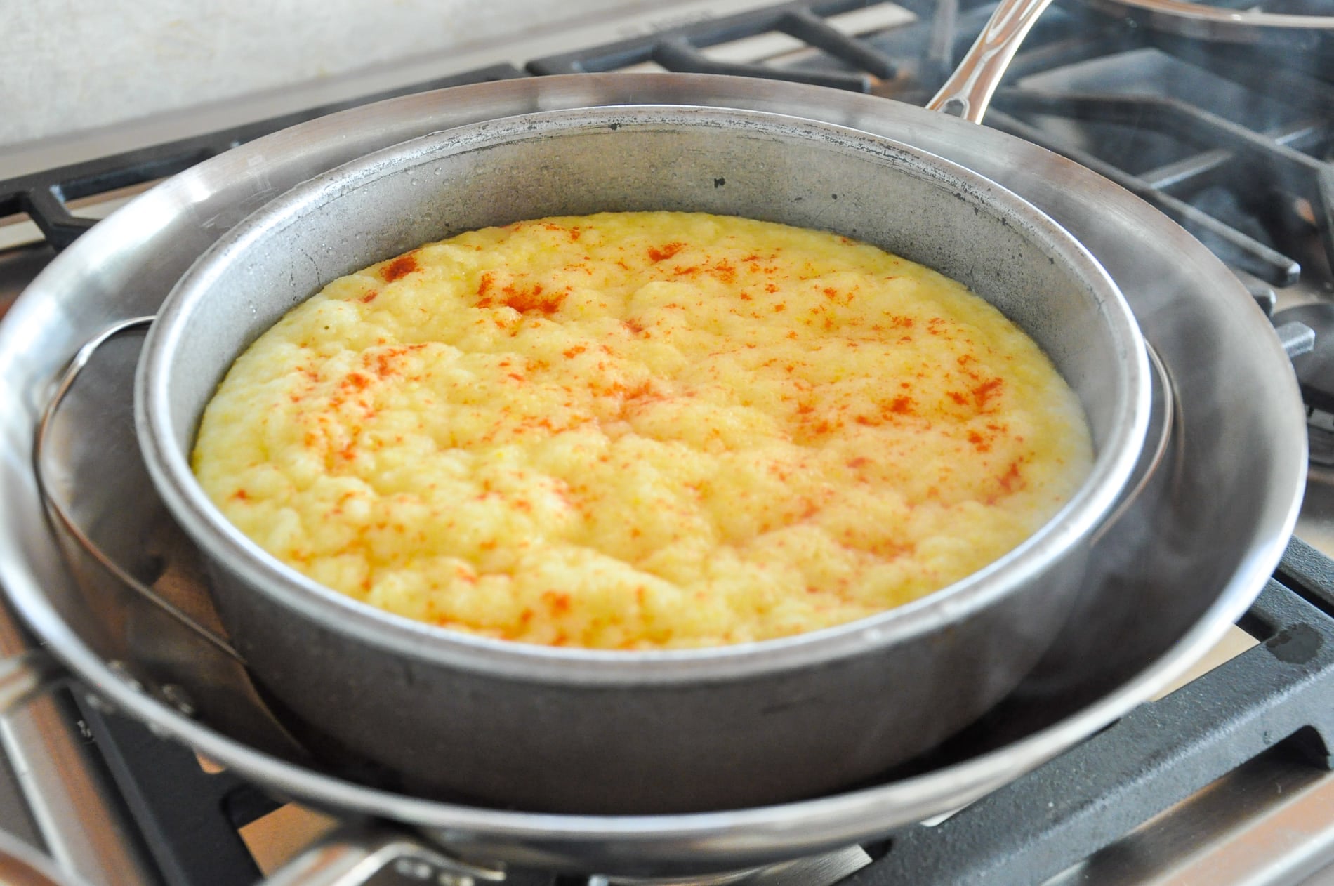A cake pan filled with steamed rava dhokla sits inside a larger pan on a stovetop, with steam rising from the setup.