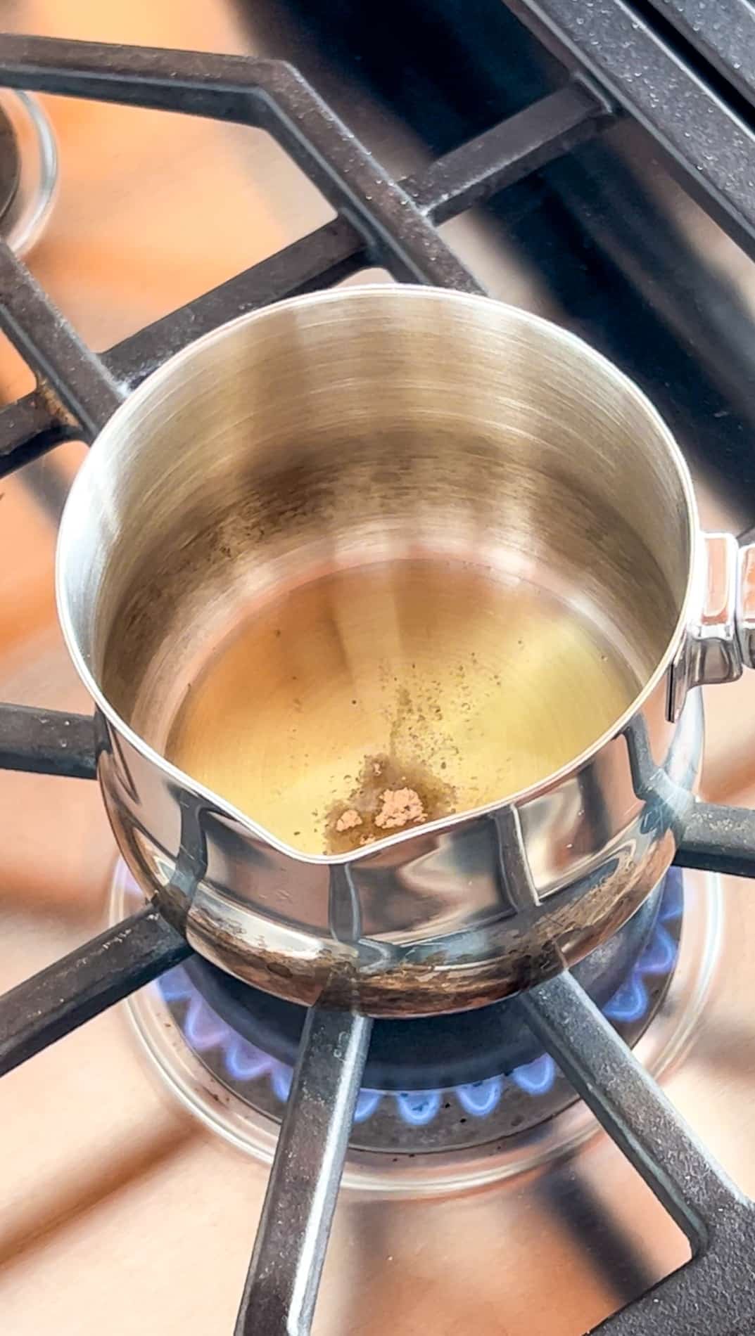 A small stainless steel pot with oil is heating on a gas stove burner. Hing (Asafoetida) has been added to the pot.