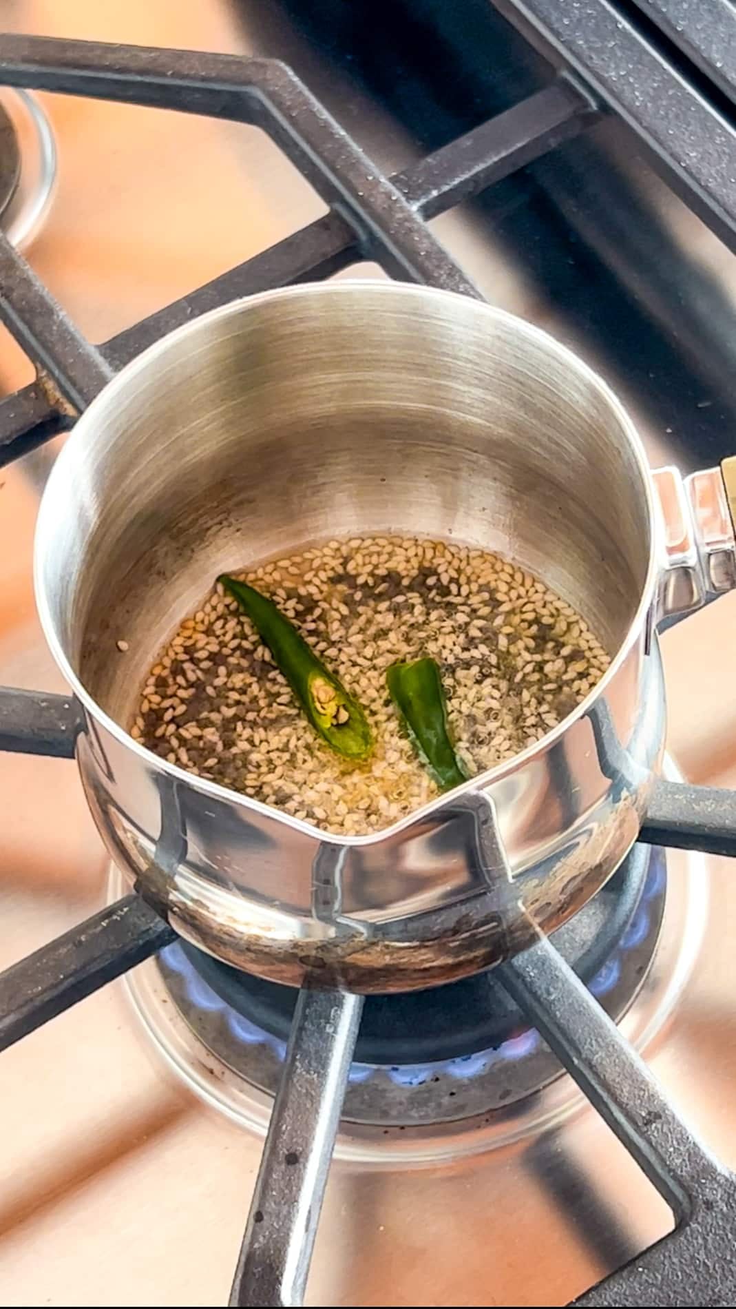 A small stainless steel pot with hot oil, cumin seeds, and two green chilies is heating on a gas stove burner.