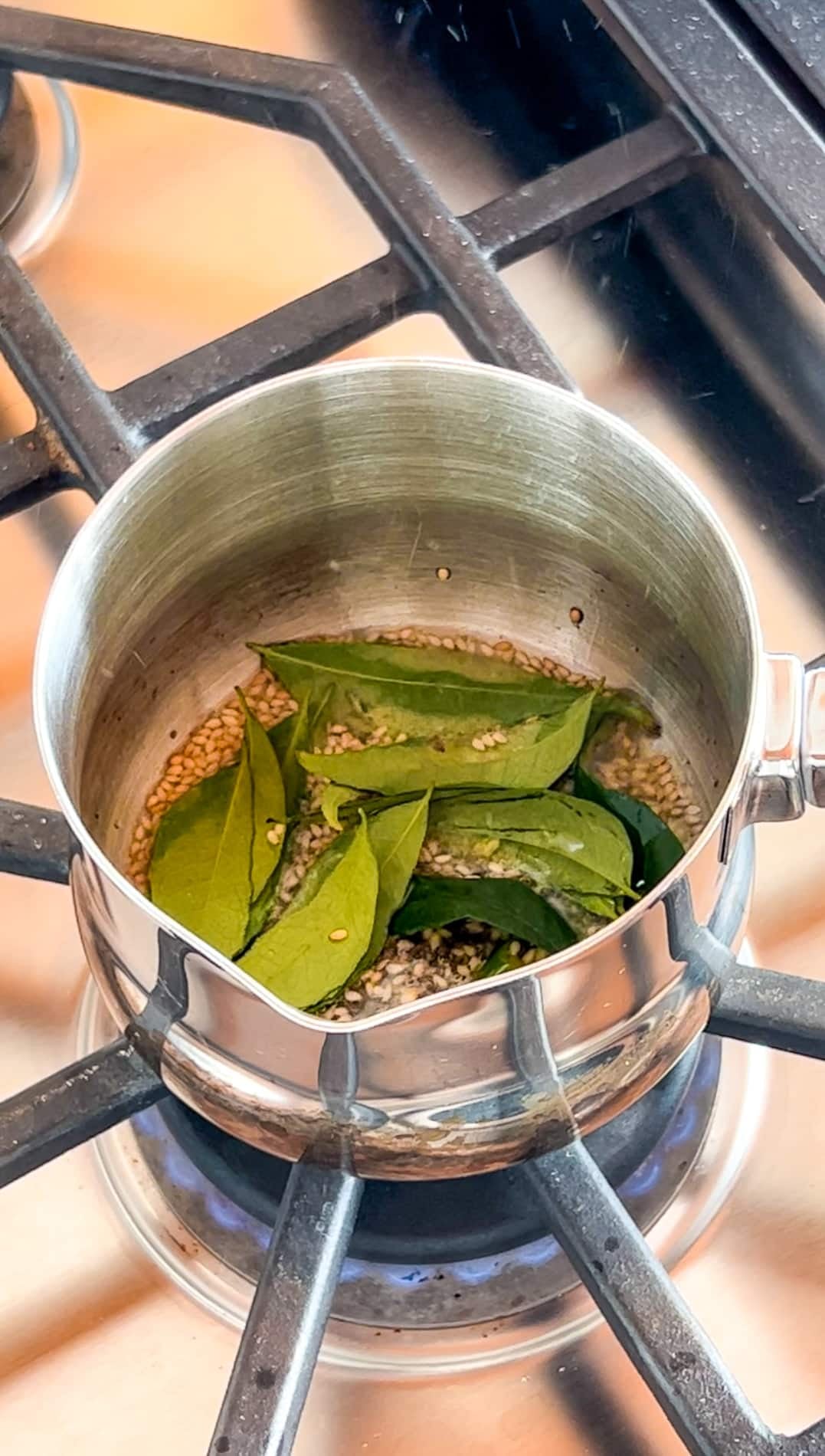 A stainless steel pot on a stove contains sesame seeds, green chili, and fresh curry leaves being heated.