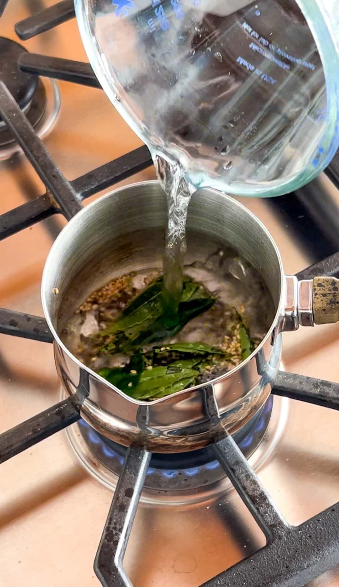 Water is being poured from a measuring cup into a saucepan with green leaves and spices on a lit stove burner.