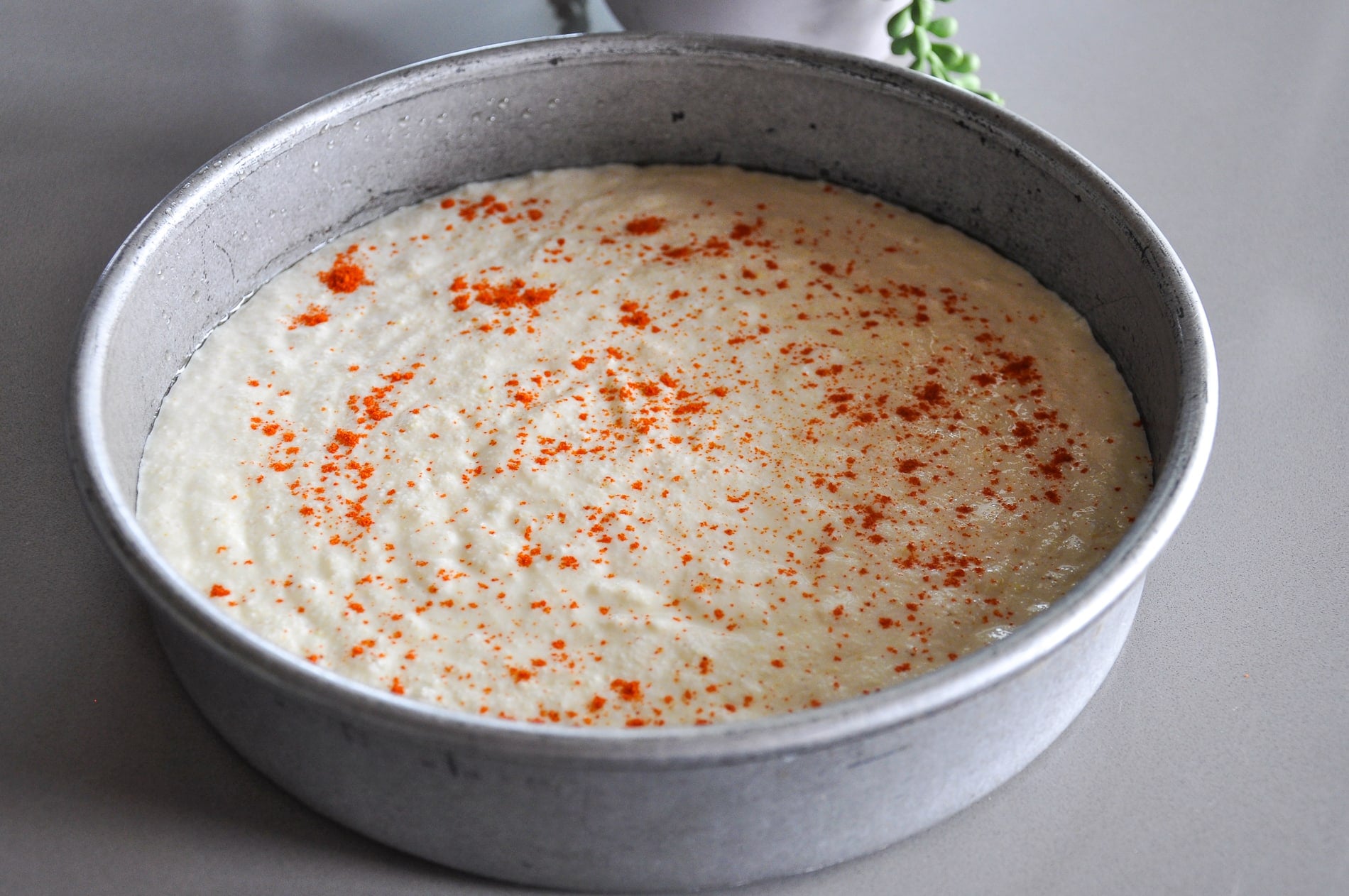 Round metal baking pan filled with uncooked dhokla batter, lightly sprinkled with red chili powder, sitting on a gray surface.