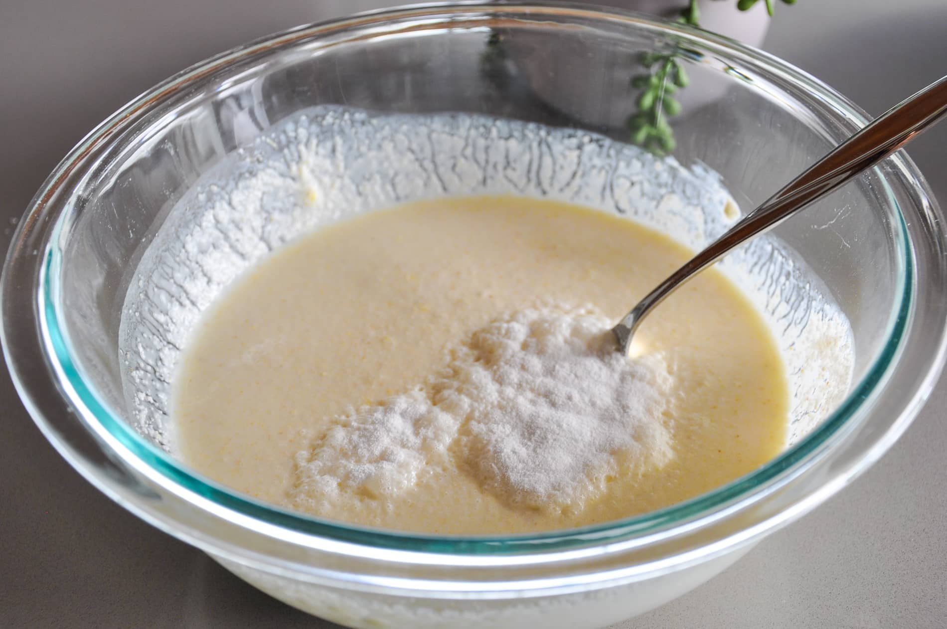 A glass bowl filled with a pale yellow dhokla batter mixture, with a spoon resting inside and ENO on the surface.