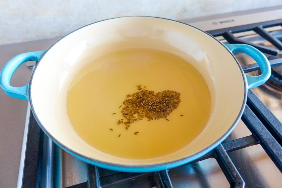 A blue-handled pan with hot oil and cumin seeds sits on a stovetop burner, with the seeds starting to sizzle.
