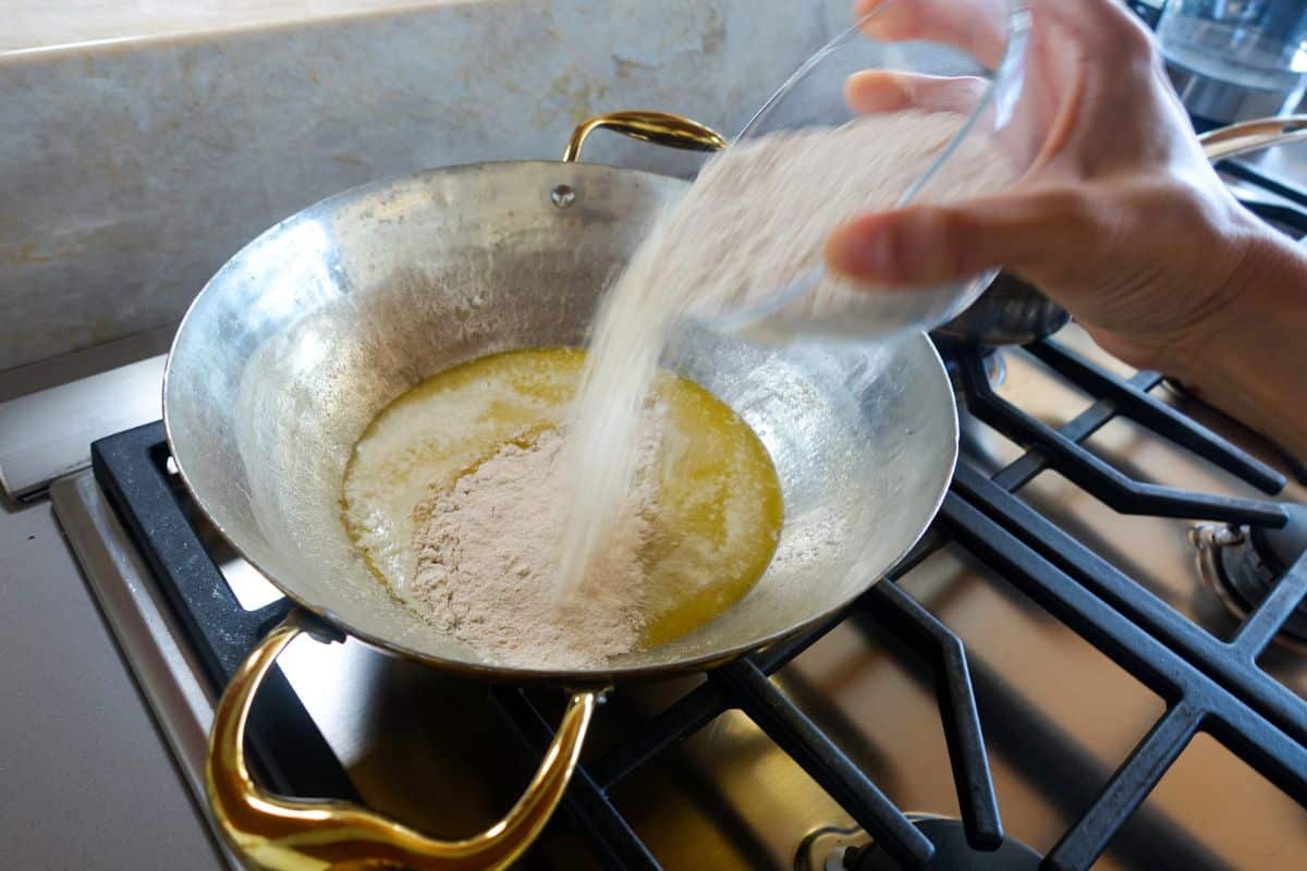 A hand pours flour from a glass bowl into a pan of melted butter on a stovetop.