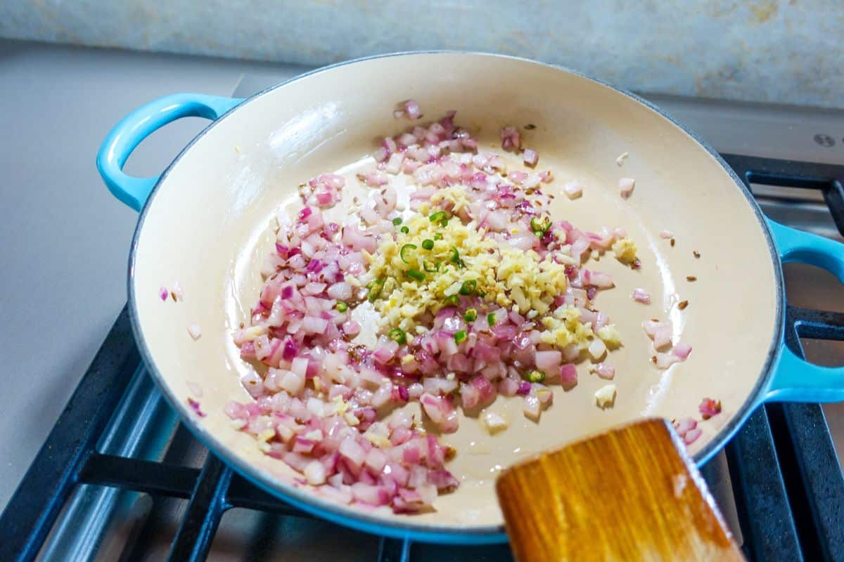 Chopped red onions, ginger, garlic, and green chili are being sautéed in oil in a light-colored pan on a stove, with a wooden spatula resting on the side.