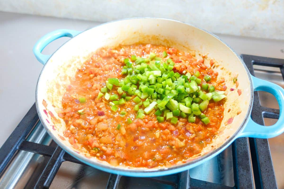 A blue-handled pan on a stove contains a tomato-based mixture with chopped green bell peppers added on top.