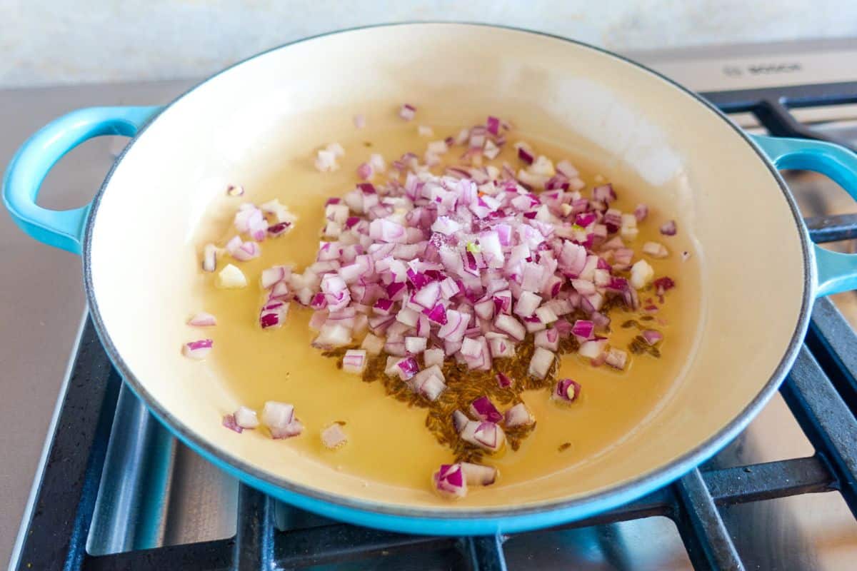 Chopped red onions and cumin seeds cooking in oil in a large blue-handled pan on a gas stove.