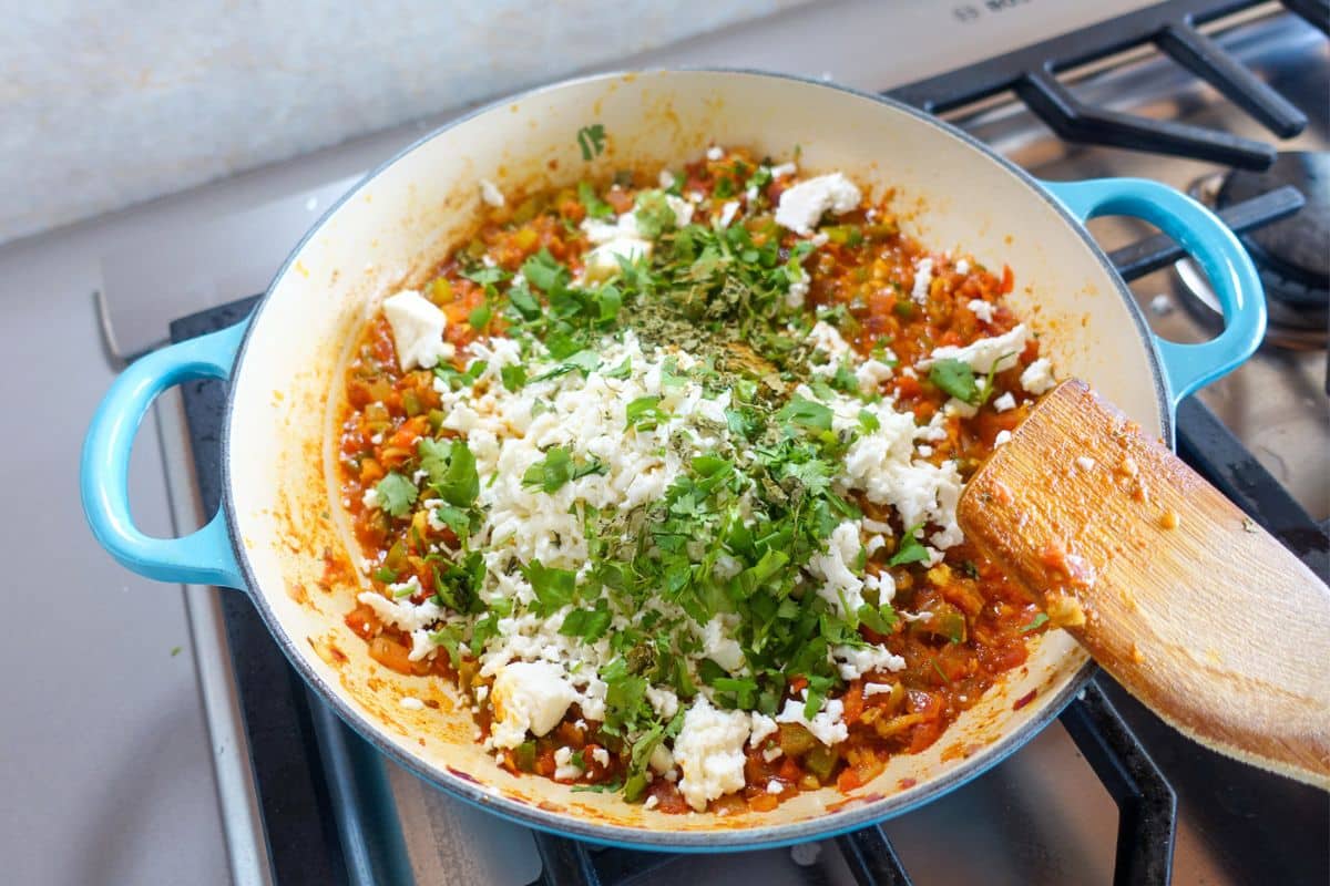 A pan of sautéed vegetables topped with chopped herbs and crumbled paneer sits on a stovetop, with a wooden spatula resting on the side.