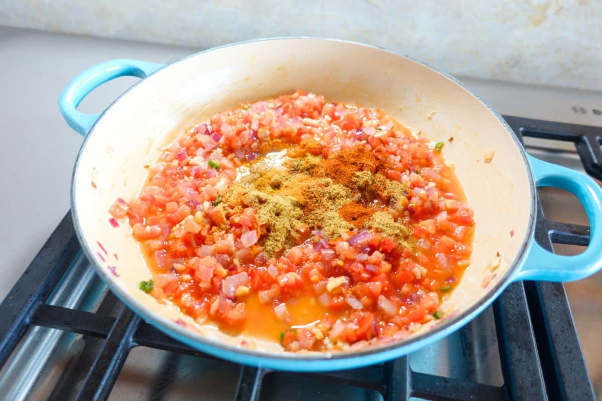 Chopped tomatoes and onions cook in a blue pan on a stove, with ground spices sprinkled on top.