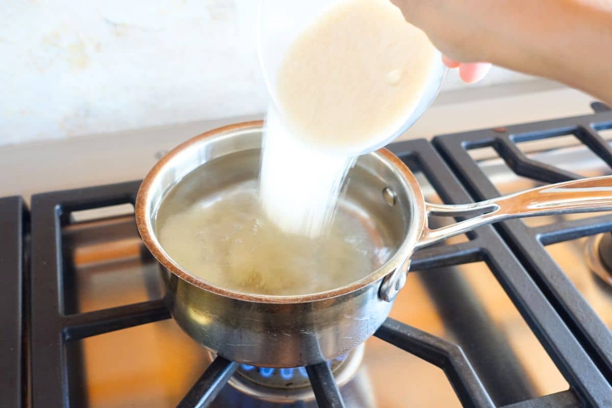 A hand pours granulated sugar from a bowl into a small pot of water on a gas stove.