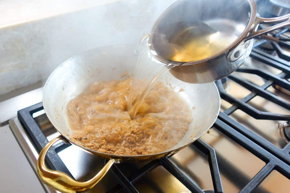 Sugar Syrup is being poured from a small pot into a pan with the roasted butter and flour mixture on a gas stove, causing steam to rise.