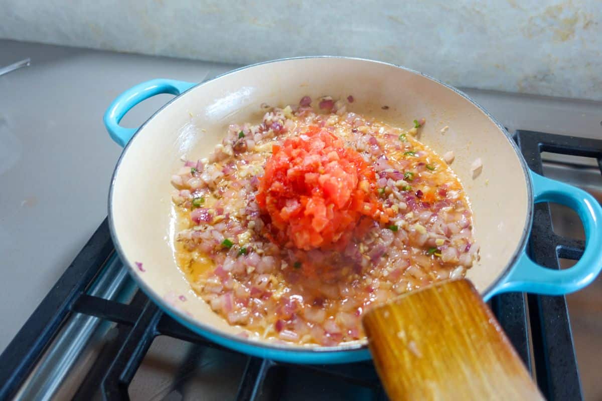 Chopped onions and tomatoes cooking in a blue pan on a stove, with a wooden spatula resting on the edge.