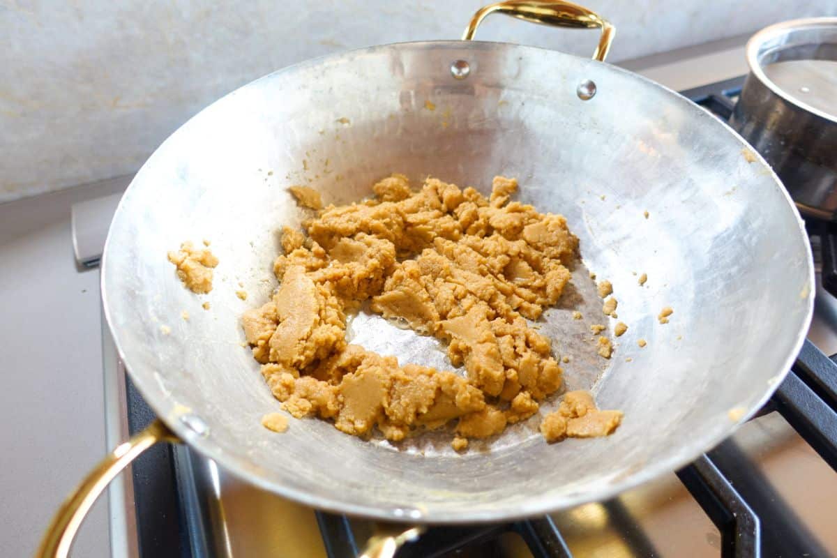 A brass pan on a stove contains a coarse, light brown mixture being cooked. A pot with water is partially visible in the background.
