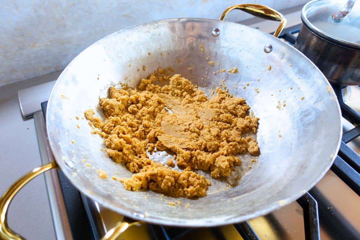A brass pan with handles contains a crumbly, golden-brown mixture, placed on a gas stove with a pot visible in the background.