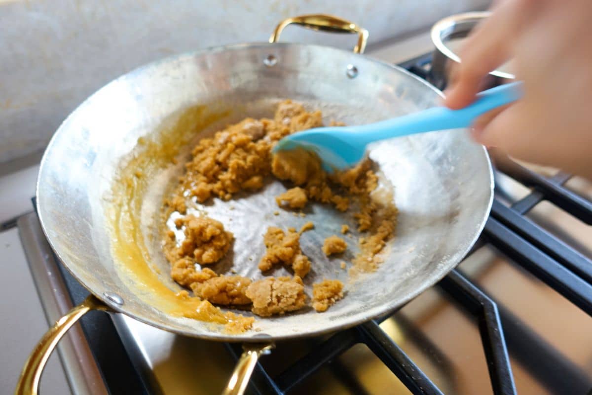 A person stirs a thick brown mixture in a brass pan on a stovetop using a blue spatula.
