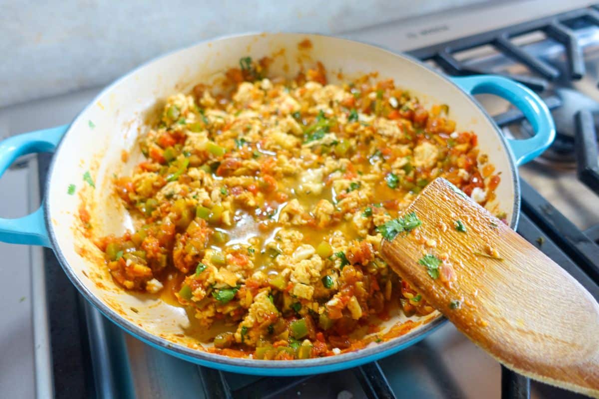 Scrambled paneer with diced vegetables and herbs cooking in a blue frying pan on a gas stove, with a wooden spatula resting on the pans edge.