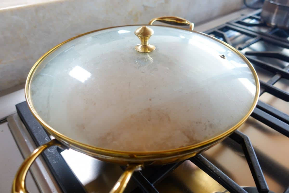 A glass-lidded brass pan sits on a gas stove burner, with condensation visible on the inside of the lid, as Atta Halwa simmers gently within.
