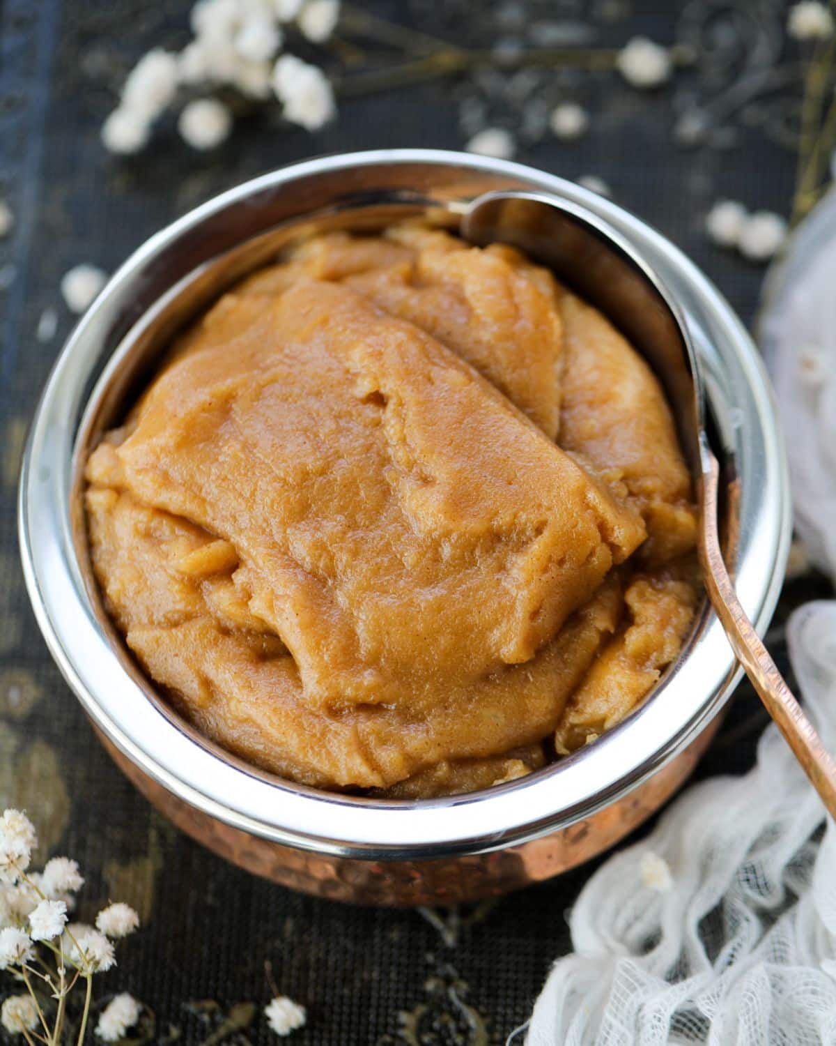 A metal bowl filled with smooth, brown atta halwa, with a spoon resting on the rim; white flowers and fabric are in the background.