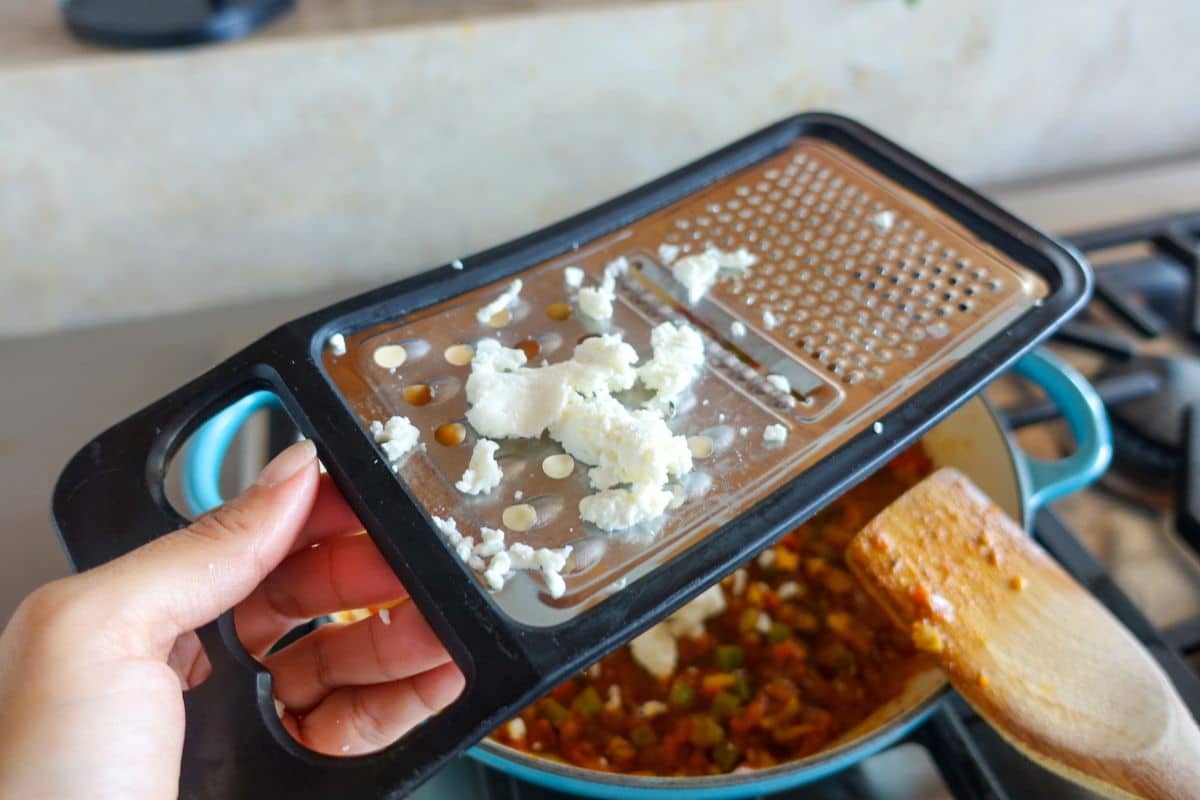 A hand holds a box grater with grated paneer above a pot of food on a stove, and a wooden spoon rests on the pots edge.