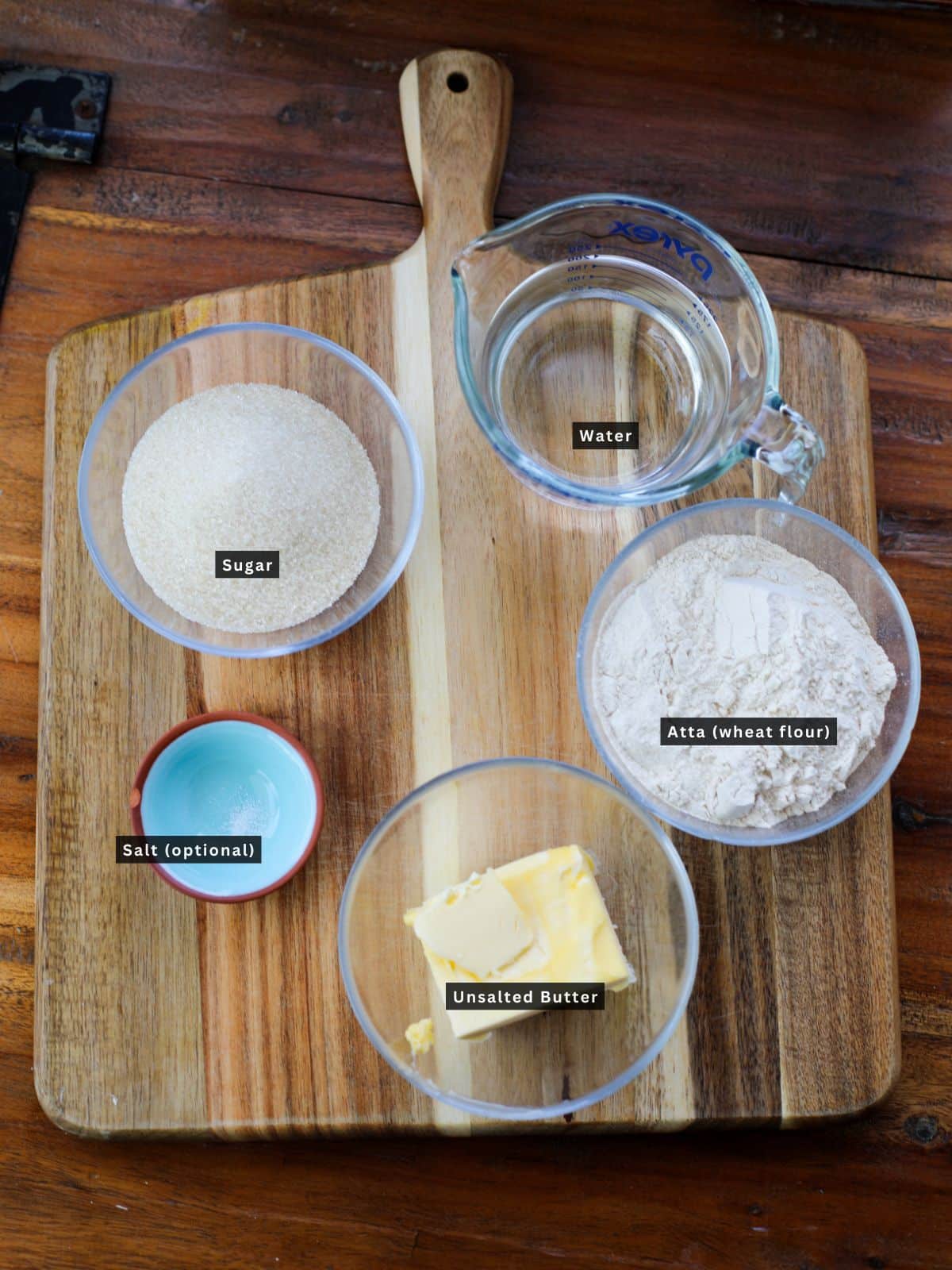 A wooden board with bowls containing sugar, wheat flour, a small dish of salt, a glass measuring cup of water, and a bowl of unsalted butter, each labeled.
