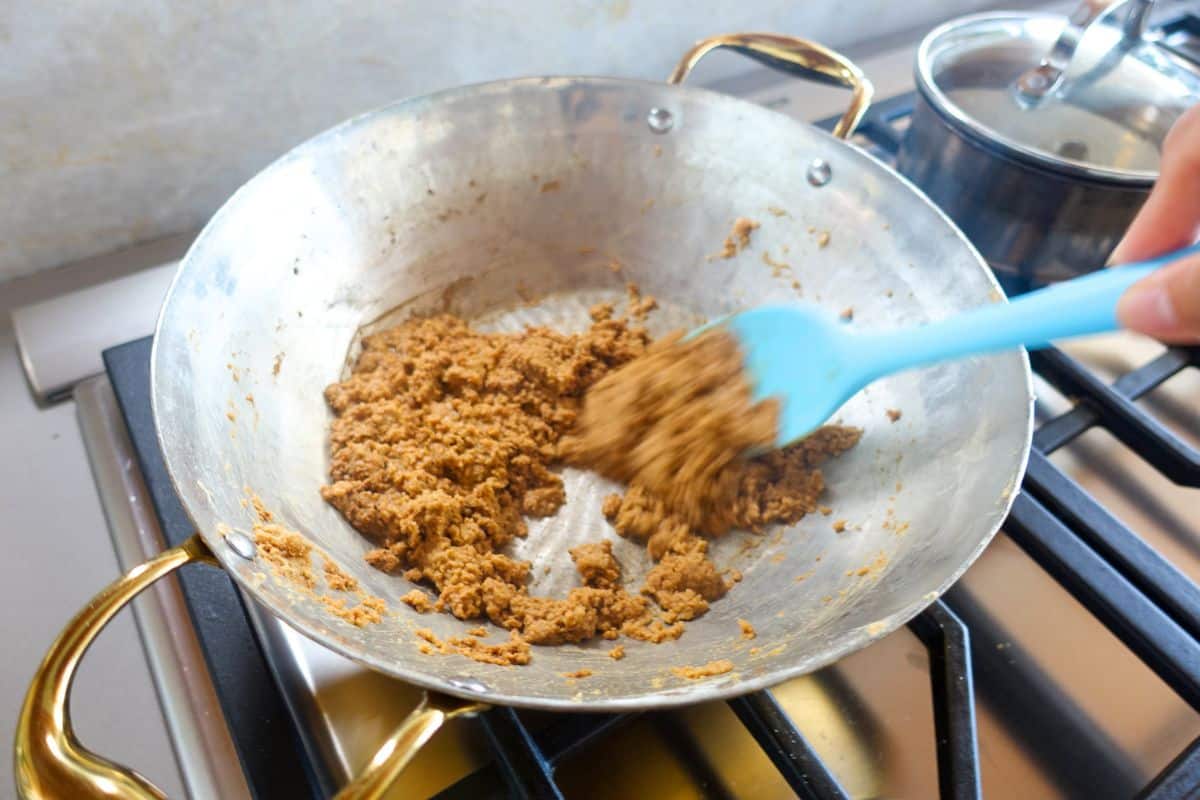A person stirs browned flour in a large brass pan with a blue spatula on a stovetop; a pot with a lid is in the background.