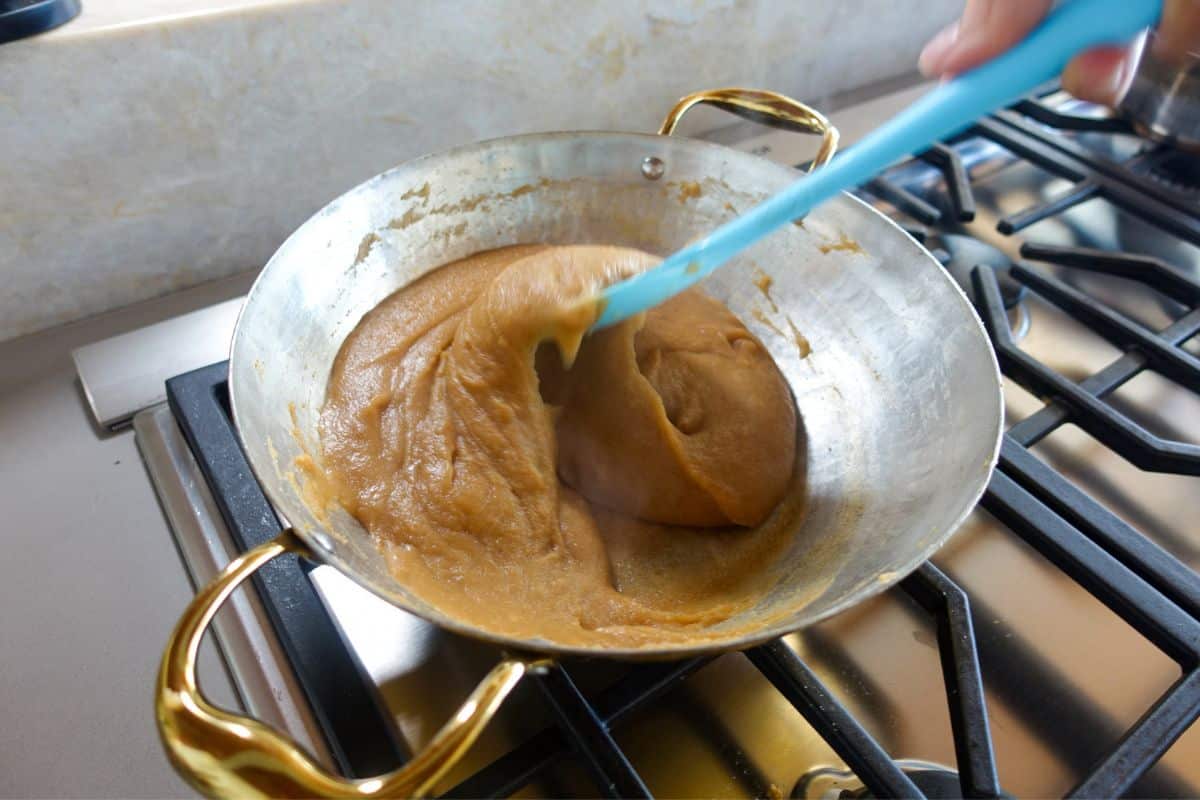 A thick, brown mixture of Kada Prasad is being stirred in a metal pan with a blue spatula on a gas stove.