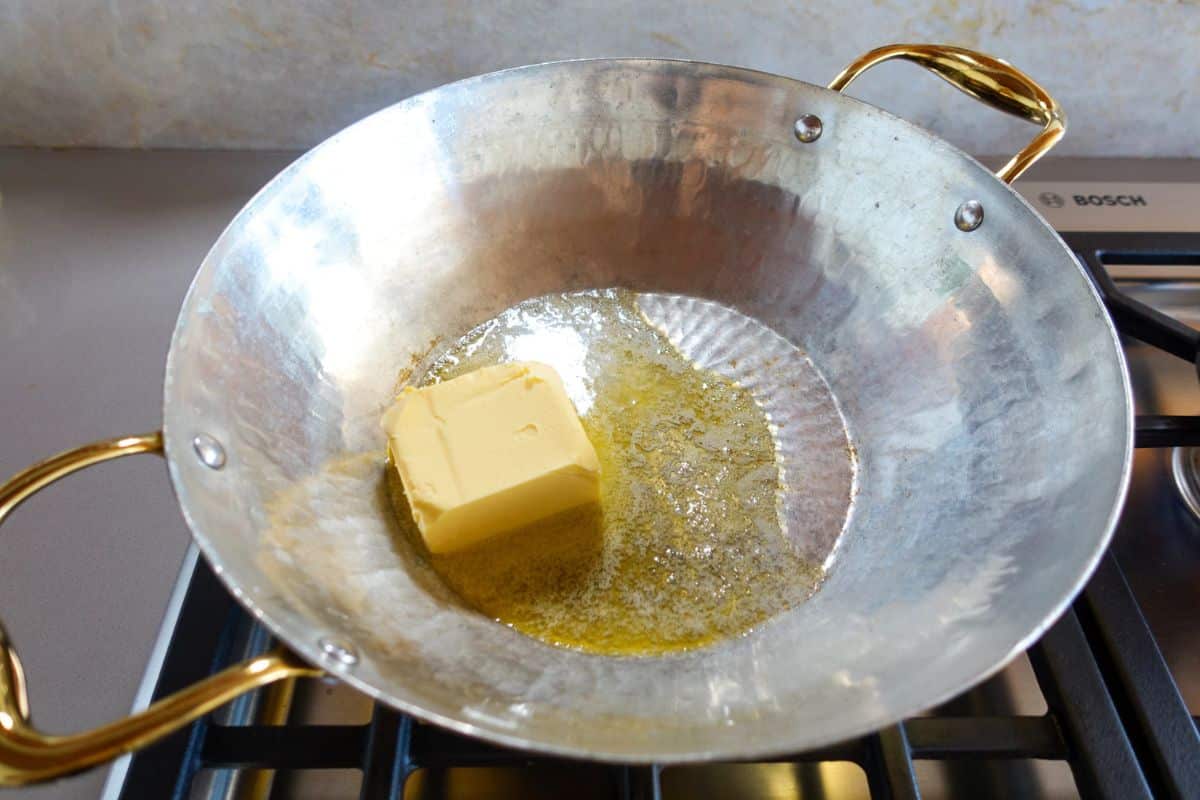 A block of butter is melting in a brass pan with gold handles on a gas stove.
