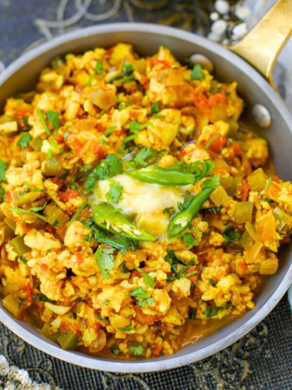 A shallow pan filled with a colorful paneer bhurji, garnished with green chilies, fresh herbs, and a small dollop of butter, placed on a patterned fabric.