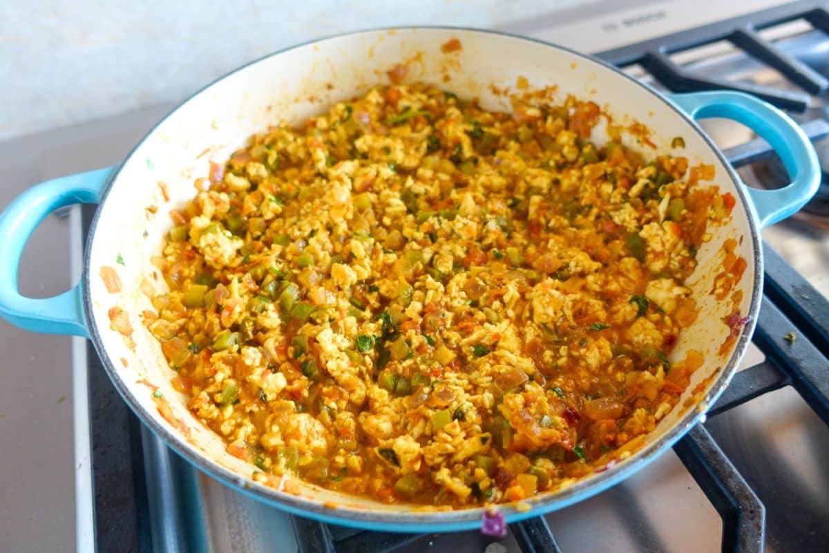 Scrambled paneer cooked with tomatoes, peppers, and spices in a blue-handled pan on a stove.