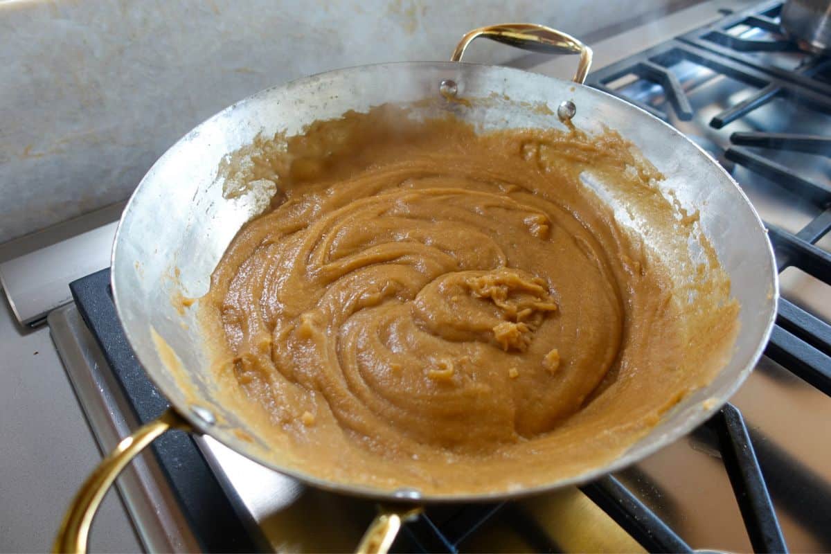 A metal pan with light brown, thick Kada Prasad batter being cooked and stirred on a stove.