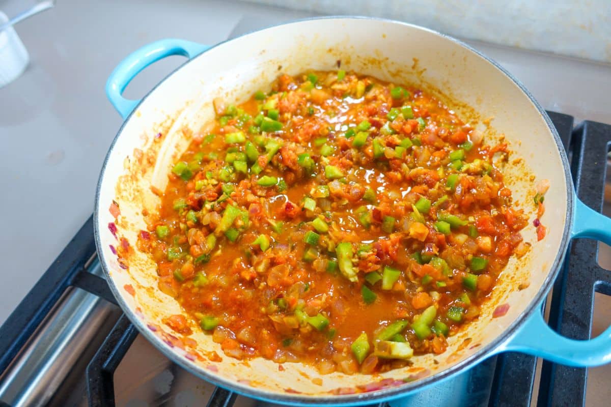 A pan on a stovetop contains a tomato-based sauce mixed with chopped green bell peppers and onions.