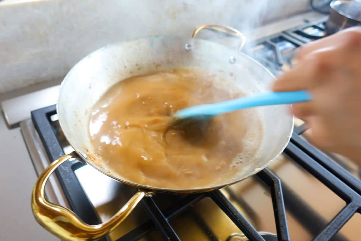 A hand stirs a brown liquid in a brass pan on a gas stove, with steam rising from the simmering atta halwa mixture.