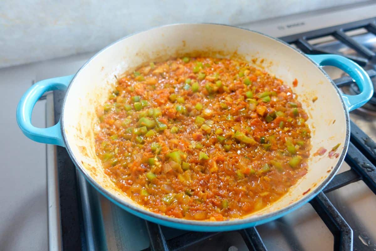 A blue-handled pan on a stove contains a simmering mixture of chopped green peppers, tomatoes, and onions in a red sauce.
