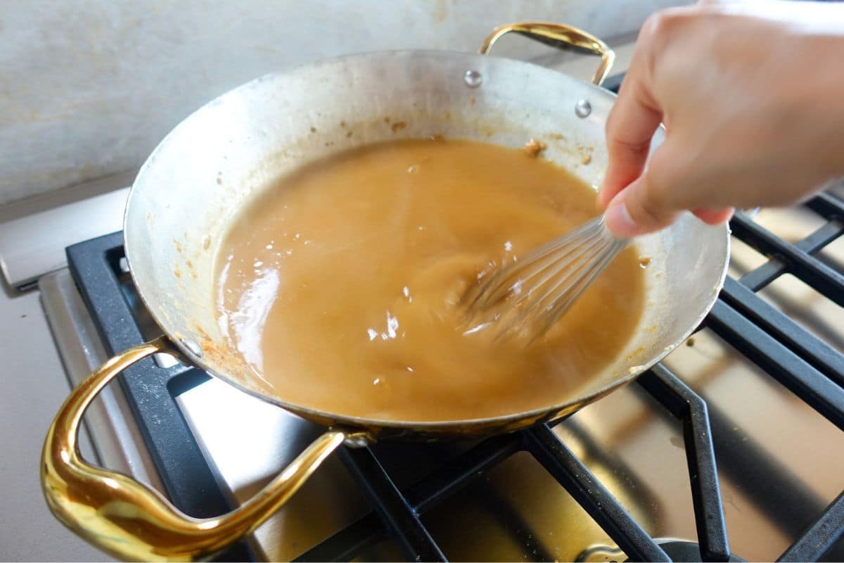 A hand uses a whisk to stir a thick brown Atta Halwa sauce in a metal pan on a stovetop.