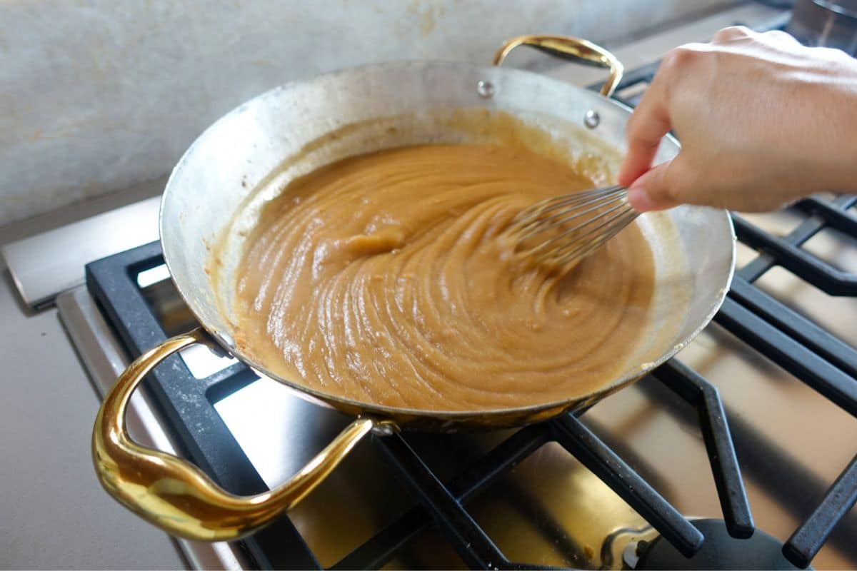 A hand uses a whisk to stir thick brown roux, similar in texture to Kada Prasad, in a metal pan with gold handles on a stovetop.