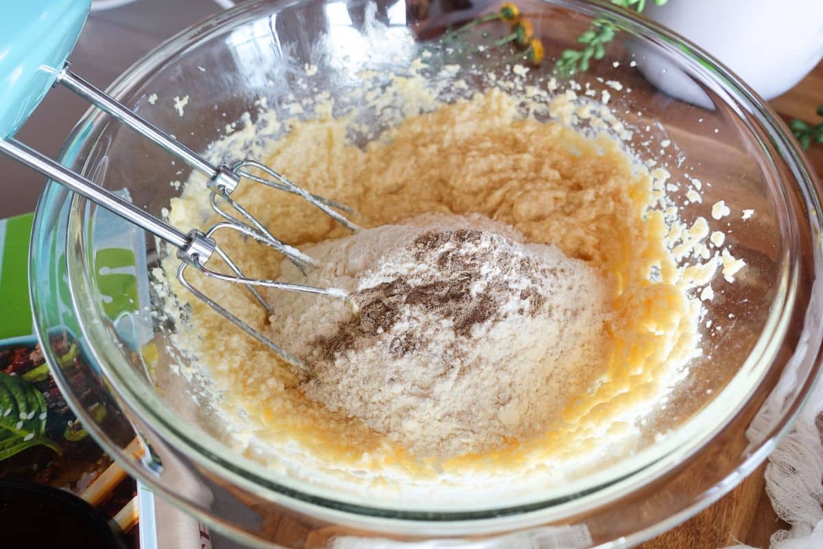 Electric hand mixer beaters mixing flour and a yellow batter in a glass bowl.