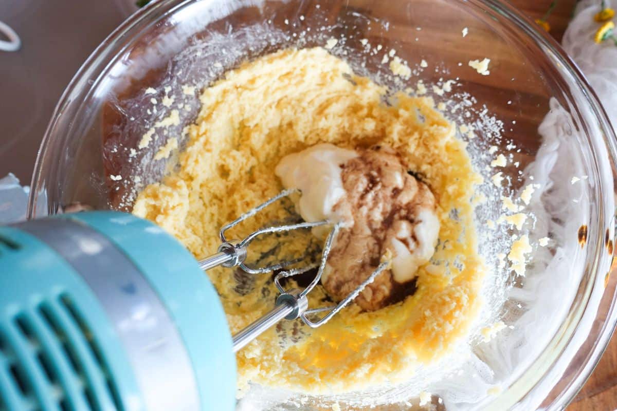A hand mixer blends a mixture of creamed butter, yogurt, and vanilla in a glass bowl.