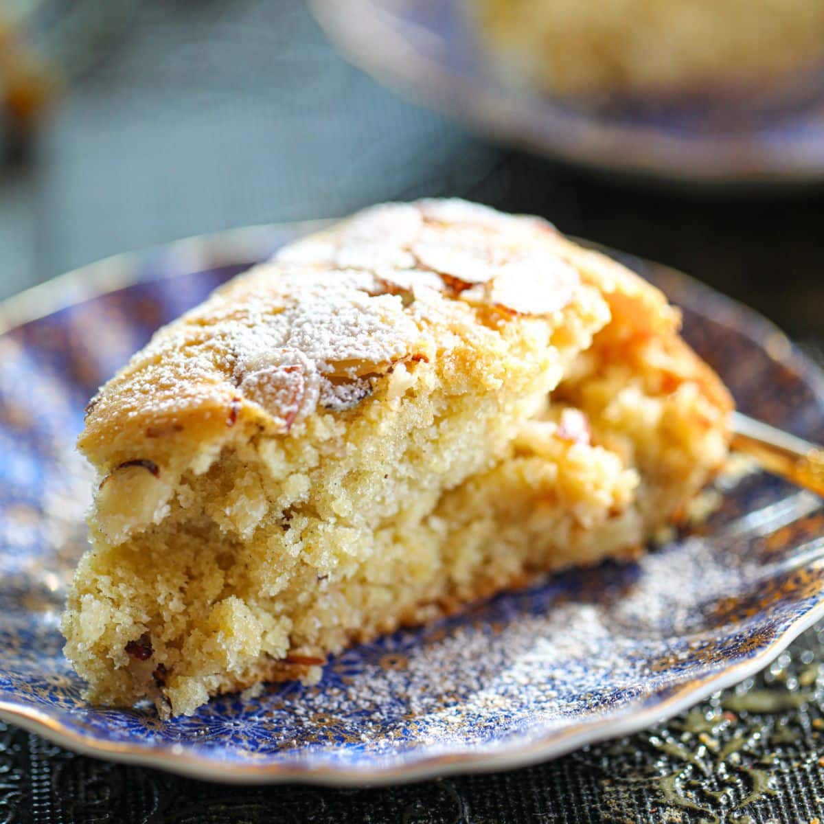 A slice of almond cake topped with powdered sugar and sliced almonds on a patterned blue plate.
