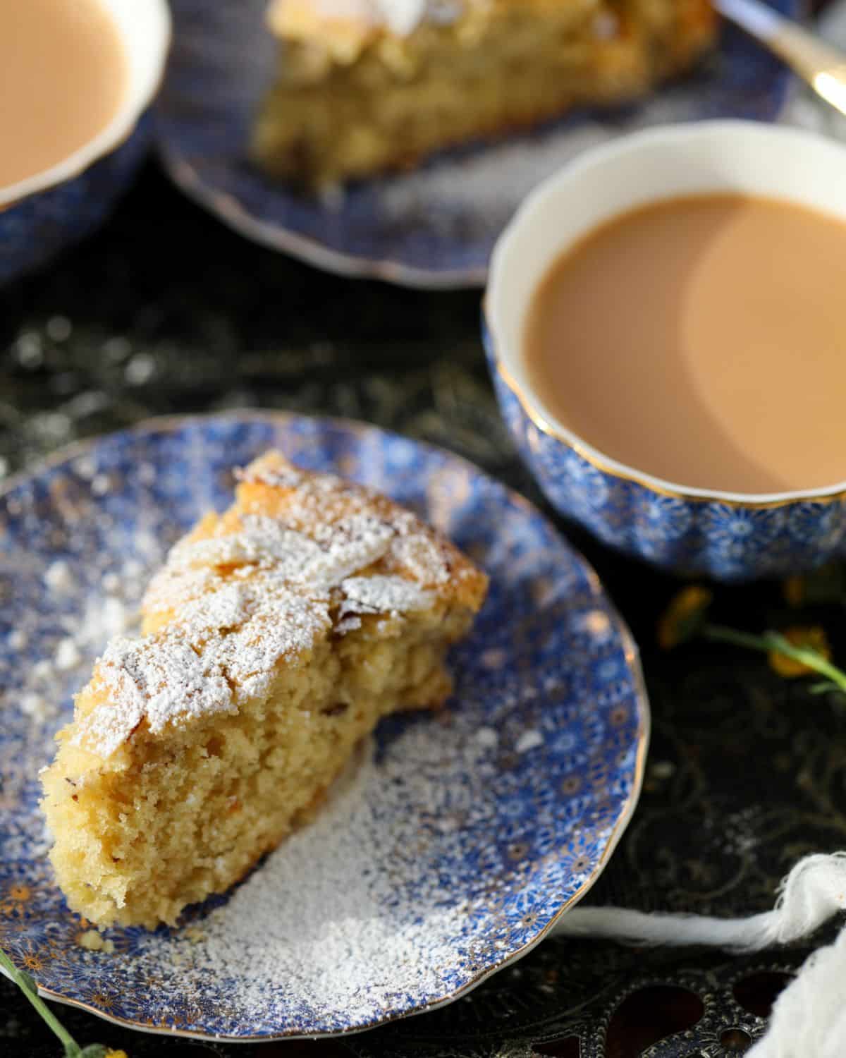 A slice of cake dusted with powdered sugar on a blue patterned plate, next to a cup of tea.