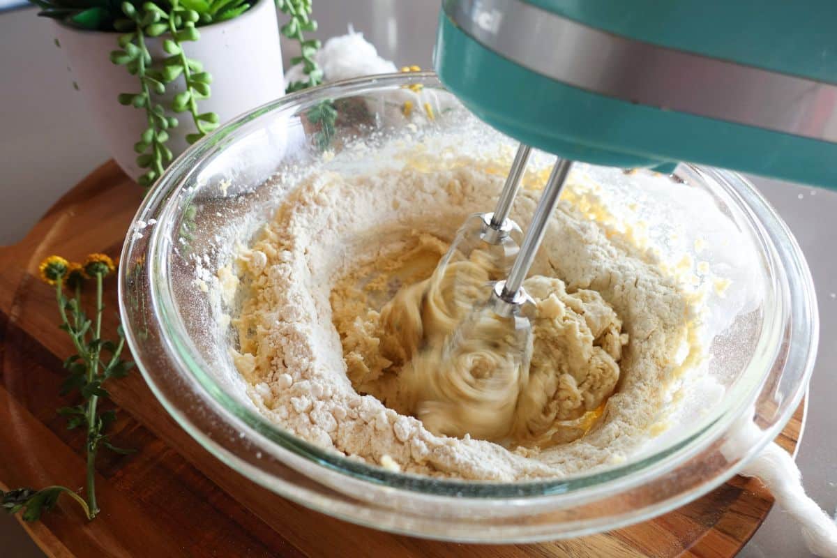 A turquoise electric hand mixer blending flour and dough in a glass mixing bowl.