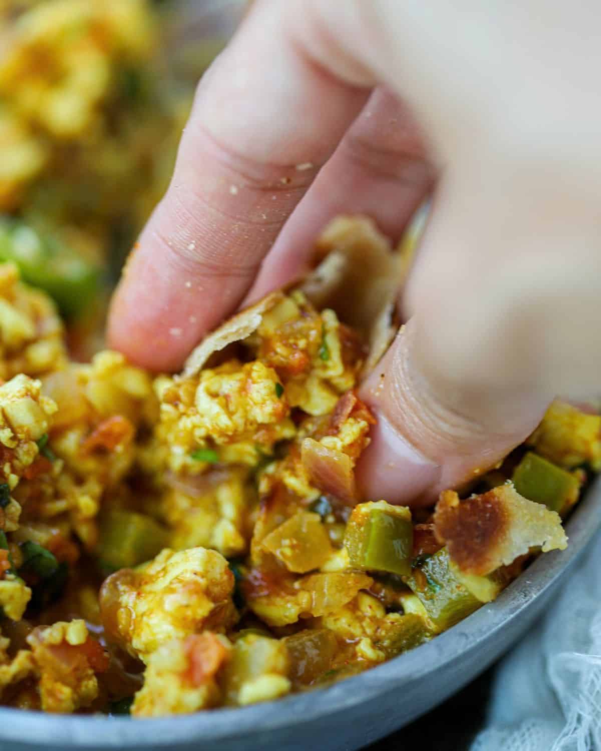 A hand uses a piece of roti to scoop up a serving of spiced Punjabi paneer bhurji from a pan.